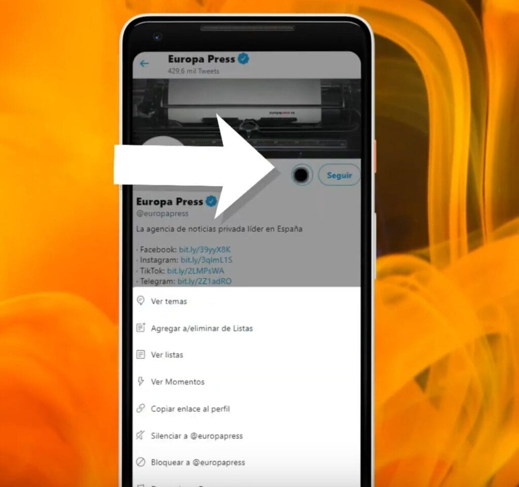 La pantalla de un smartphone muestra la aplicación de Twitter con una página de perfil de "Europa Press". Una flecha blanca resalta el botón "Seguir". El fondo presenta un diseño abstracto en amarillo y naranja, que sutilmente sugiere tutoriales como "Cómo bloquear un usuario en Twitter".