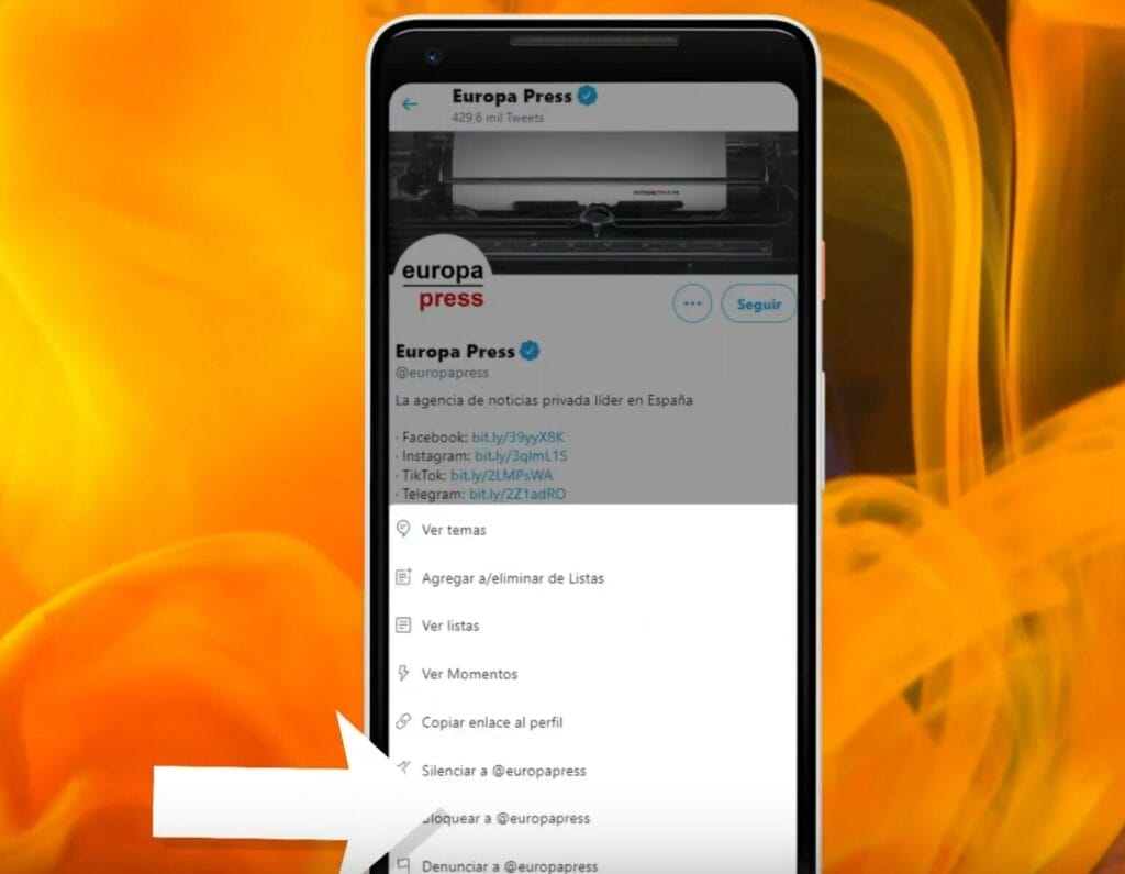 como bloquear a alguien en twitter -- La pantalla de un teléfono inteligente muestra un perfil de Twitter de "Europa Press" con opciones, entre ellas "Silenciar a @europapress", resaltada con una flecha blanca. El fondo es un patrón naranja-amarillo intenso que ilustra los pasos para bloquear un usuario en Twitter.